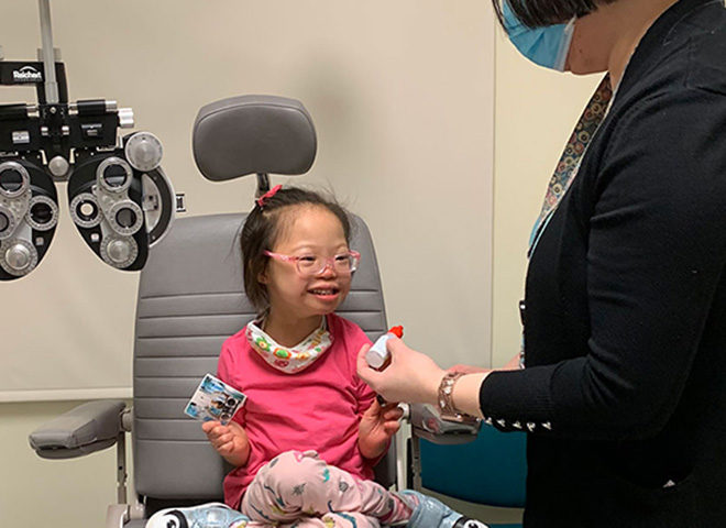 Young child in optometry chair with glasses smiling toward parent.