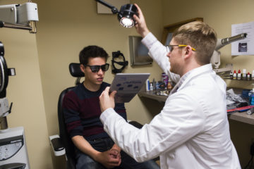 Optometrist in white coat is giving an eye exam to a male patient in an exam chair.